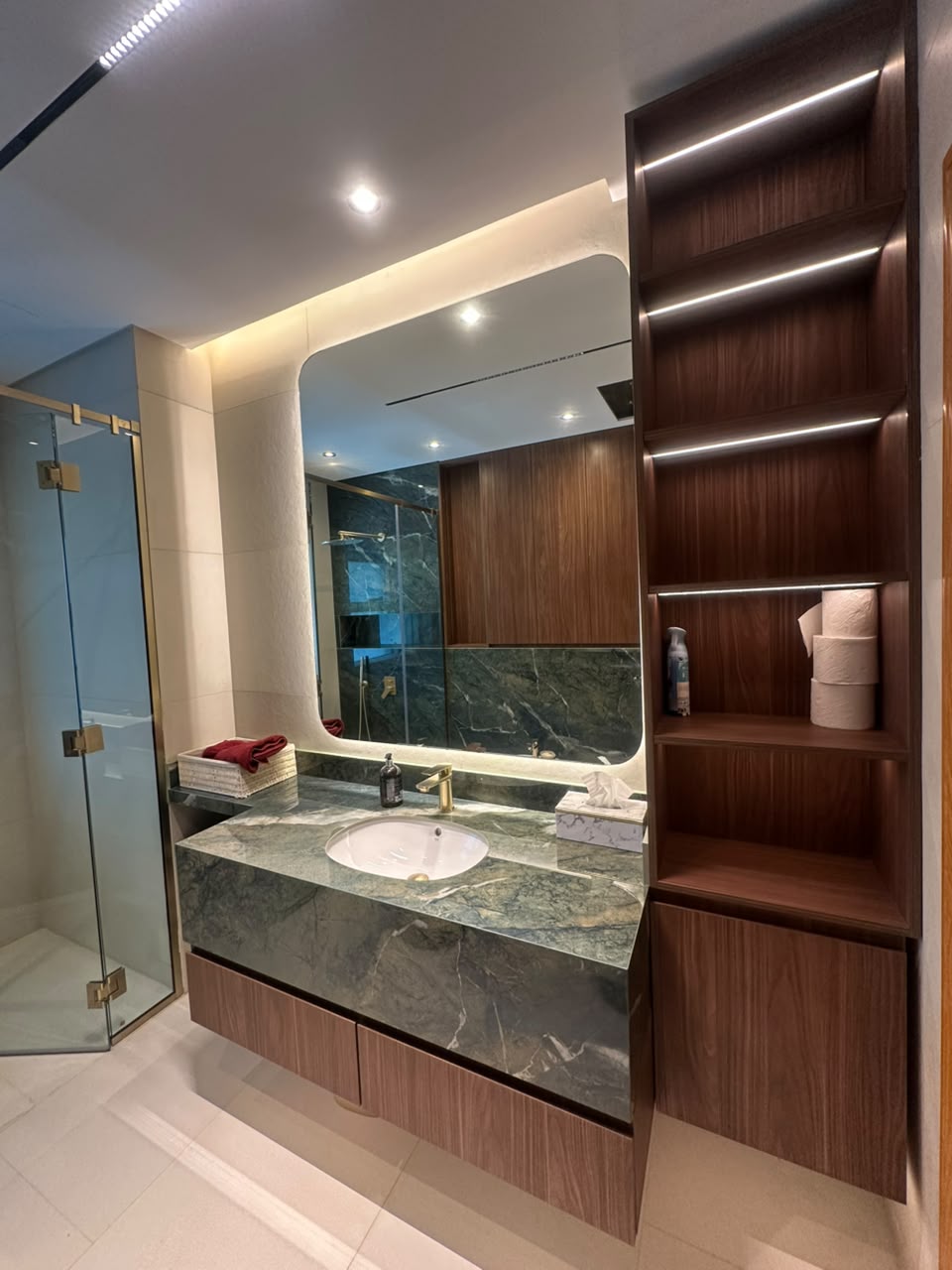 Bathroom with a green marble vanity on walnut drawers, a backlit rounded mirror and LED-lit walnut shelving